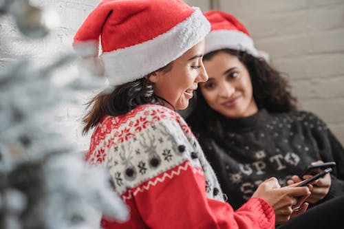 Side view of young smiling Hispanic female in Santa hat browsing mobile phone while celebrating Christmas holiday with female friend