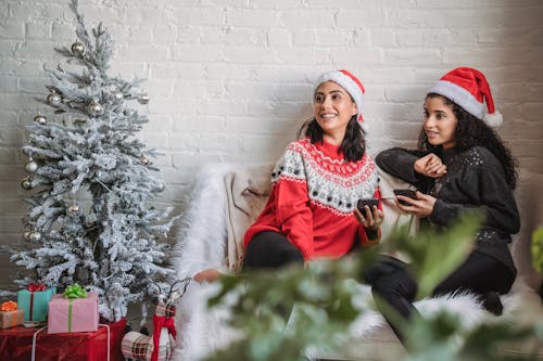 Happy young Hispanic women in Santa hat with mobile phones sitting on small sofa near decorated Christmas tree and looking away