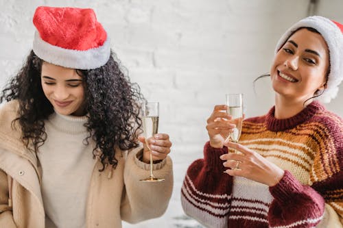 Free Positive ethnic women in warm sweaters laughing together while drinking champagne during New Year holidays Stock Photo