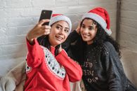 High angle of ethnic best friends in Santa hats smiling widely while taking photo on cellphone during New Year holidays