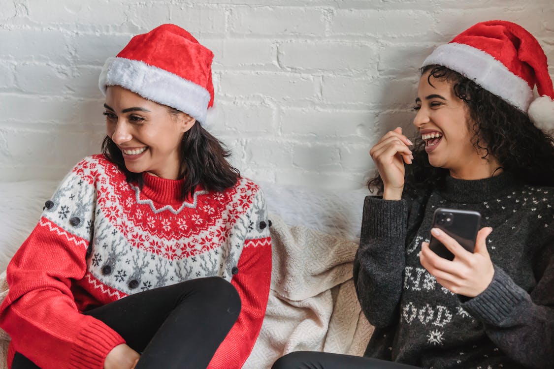 Positive ethnic female friends in warm sweaters and Santa hats having fun while using smartphone at Christmas holidays
