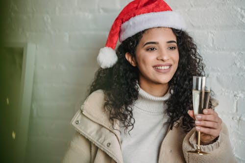 Happy ethnic female with curly hair in Santa hat smiling brightly while drinking beverage for Christmas celebration