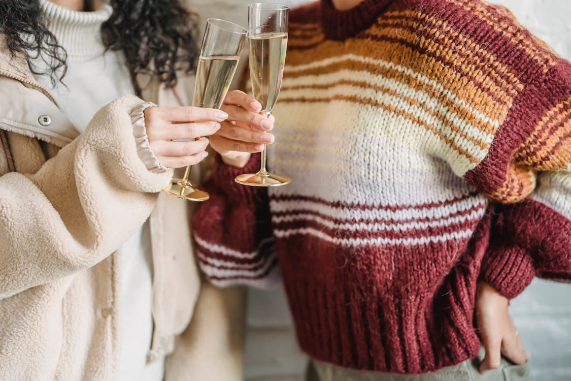 Crop faceless female friends in knitted sweaters clinking wineglasses of champagne during celebration together