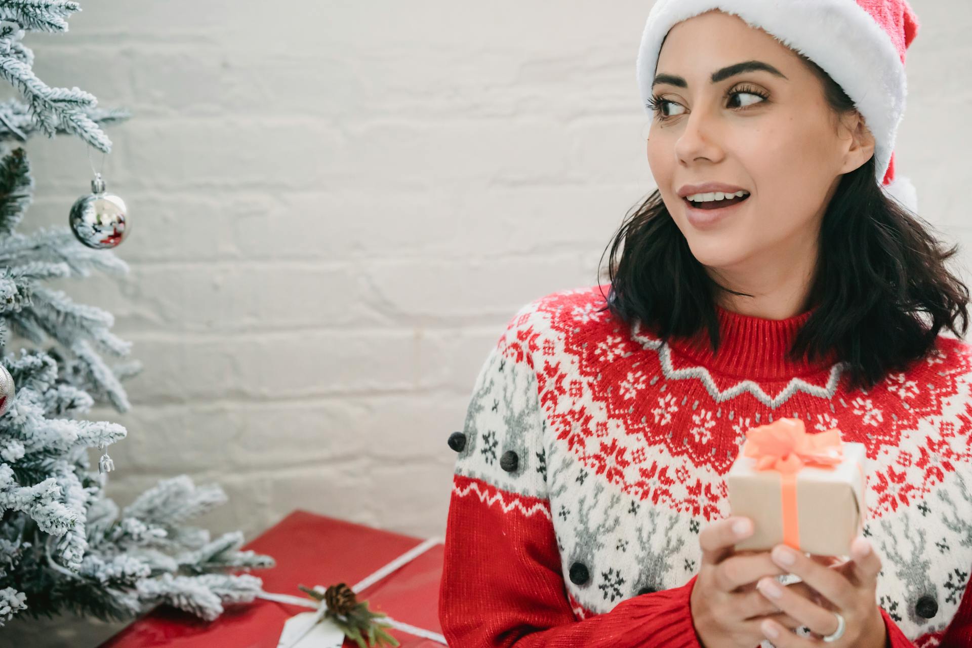 Surprised ethnic female with mouth opened in Santa hat sitting near decorated Christmas tree during presents exchange