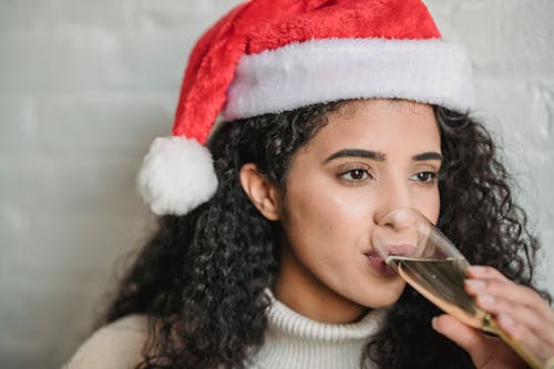 Content young ethnic brunette wearing red Santa hat and drinking sparkling champagne against white wall during Christmas celebration