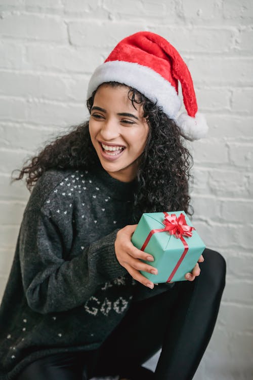 Carefree young ethnic female in cozy sweater and red Santa hat holding wrapped present and looking away in excitement during Christmas celebration