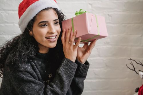 Free Smiling young ethnic female with curly hair wearing Santa hat and holding wrapped gift box near ear while looking away in excitement during Christmas party Stock Photo