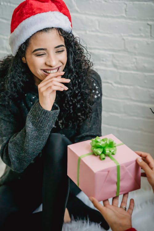 High angle of happy ethnic female in Santa hat receiving box with present for Christmas from girlfriend
