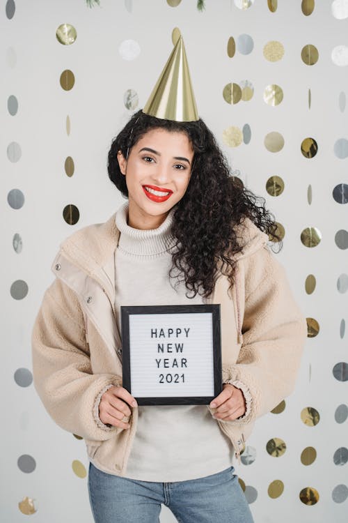 Positive ethnic female showing fame with Happy New Year 2021 text while celebrating holidays and standing against round shiny decorations