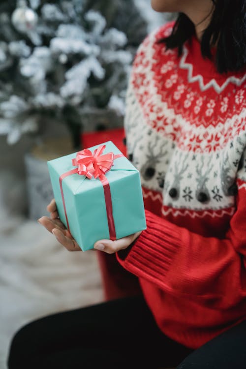 Crop anonymous female wearing knitted sweater sitting with box wrapped in gift paper and tied with ribbon near spruce branches during New Year celebration