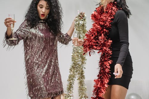 Female friends in tinsel drinking champagne and having fun while celebrating New Year against gray background