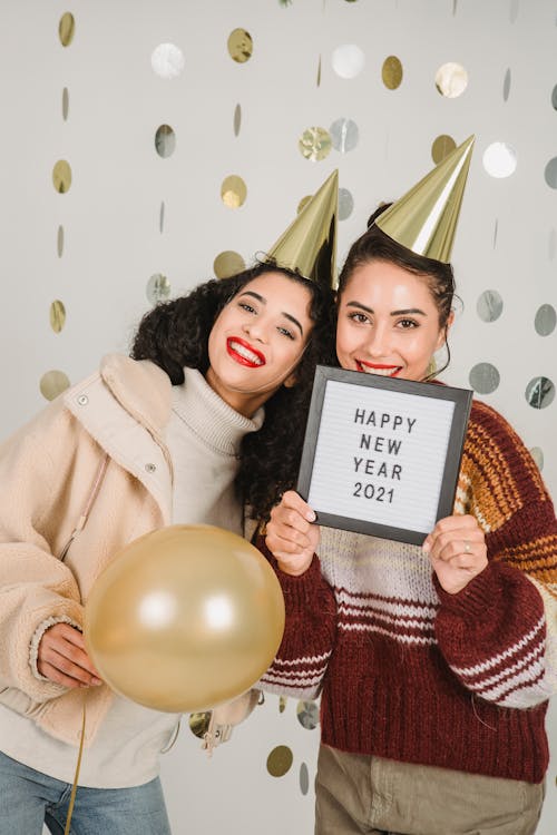 Happy ethnic best friends in cone caps standing close with framed phrase Happy New Year 2021 and balloon