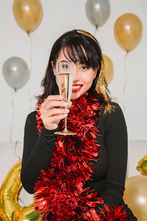 Cheerful female in red shimmering tinsel showing glass of champagne and smiling while looking at camera among balloons