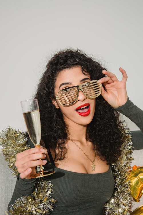 Young female with red lips and mouth opened in striped glasses and golden tinsel looking away on gray background