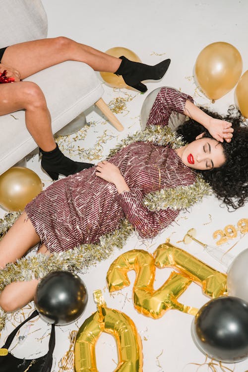 From above of young female with eyes closed in festive dress lying on floor among shiny golden and black balloons
