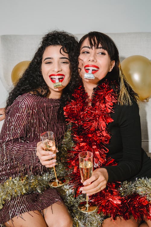 Happy friends with red lips in festive dresses and red and golden sparkling tinsel with glasses of champagne among balloons
