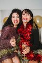 Happy friends with red lips in festive dresses and red and golden sparkling tinsel with glasses of champagne among balloons