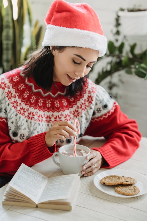 Happy smiling female in red Santa hat with cup of coffee with marshmallows and candy cane near plate with cookies