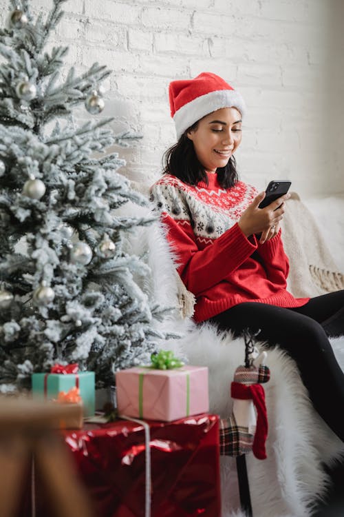 Free Delighted woman in Santa hat smiling and using smartphone Stock Photo