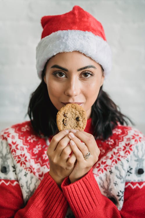 Free Happy young female in warm sweater and Santa hat eating aromatic yummy chocolate chip cookie and looking at camera during Christmas holidays Stock Photo