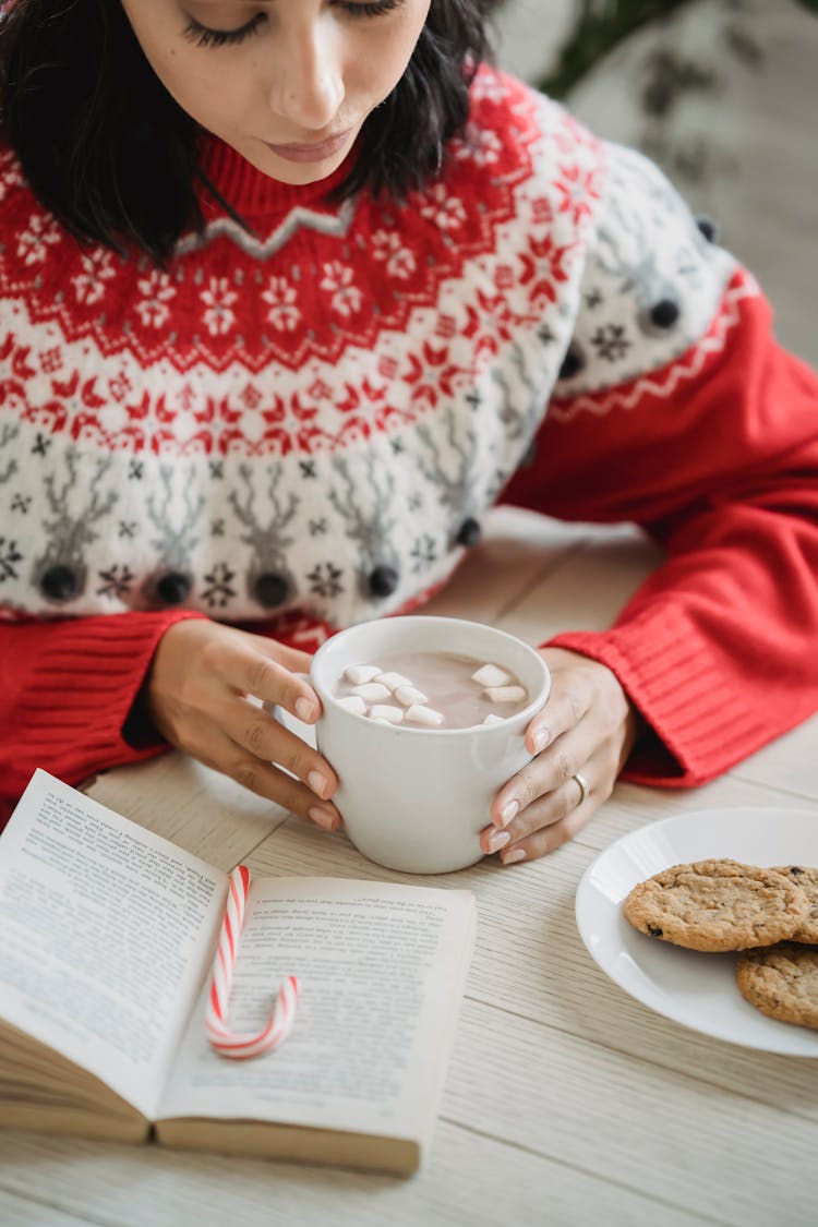 Crop Woman Enjoying Hot Chocolate And Reading Book At Table During Christmas