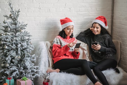 Cheerful ethnic women browsing smartphones on sofa near Christmas tree