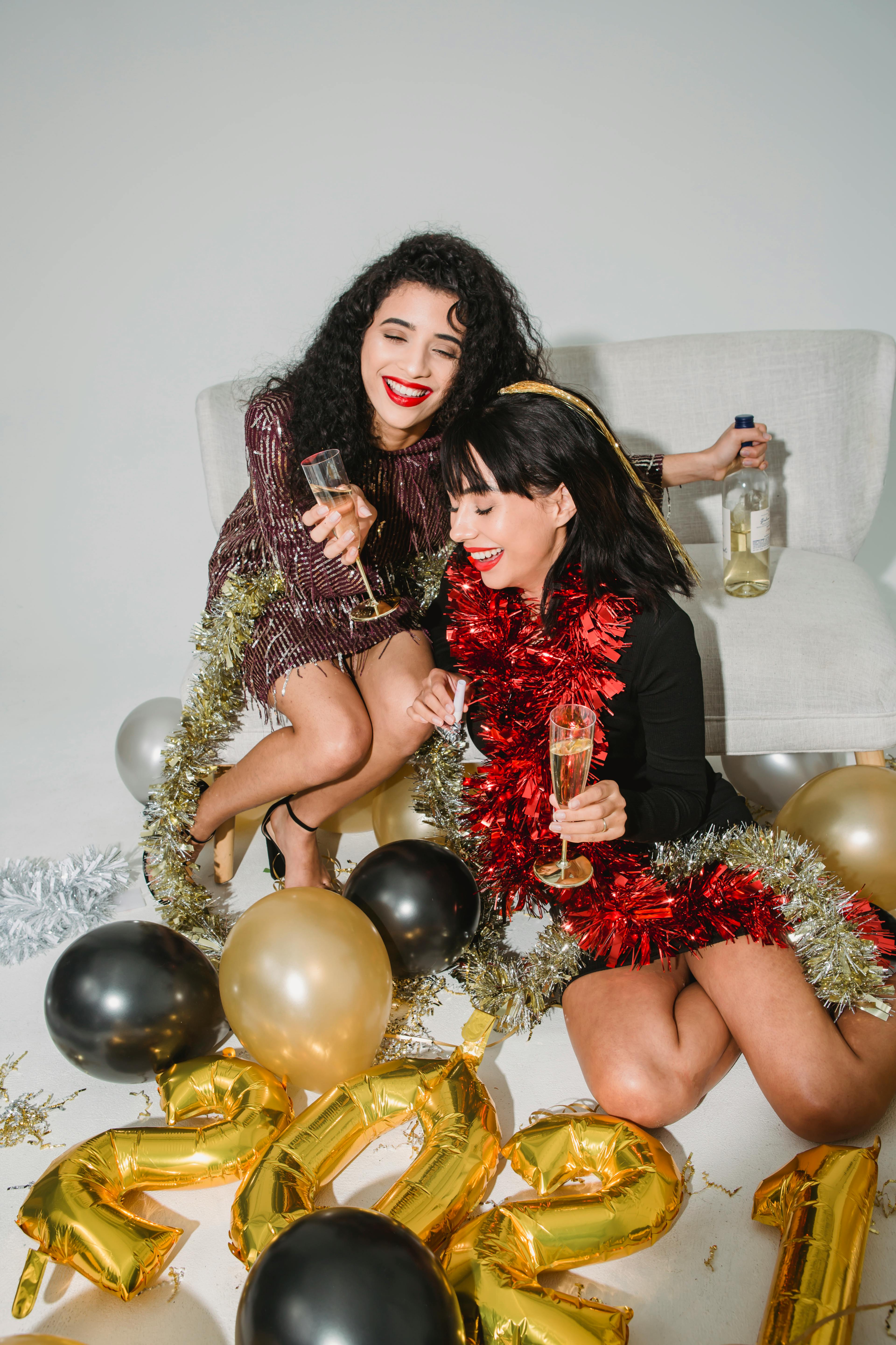 Laughing young females wearing stylish mini dresses and shiny tinsels on neck drinking sparkling champagne while sitting on floor near balloons during new year celebration party