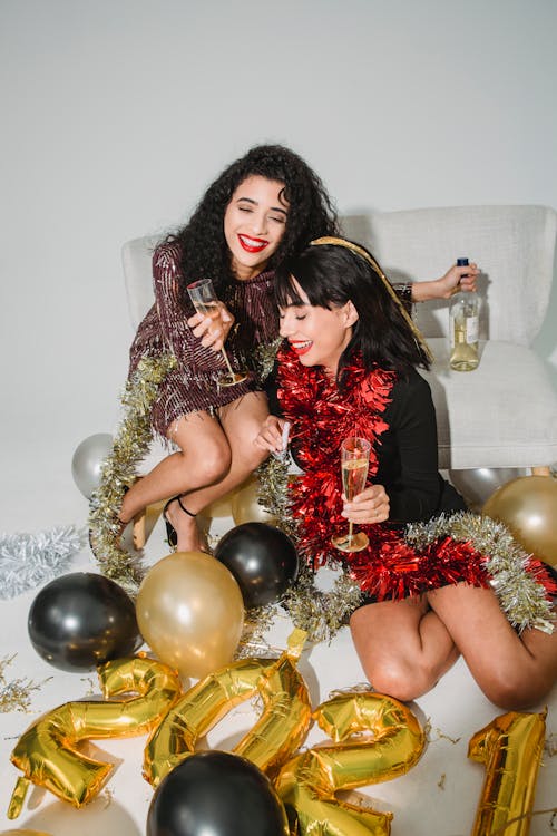 Free Laughing young females wearing stylish mini dresses and shiny tinsels on neck drinking sparkling champagne while sitting on floor near balloons during new year celebration party Stock Photo