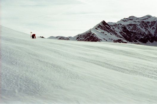 Anonymous unrecognizable person with skis travelling among snow near rocky rough slope under cloudy sky