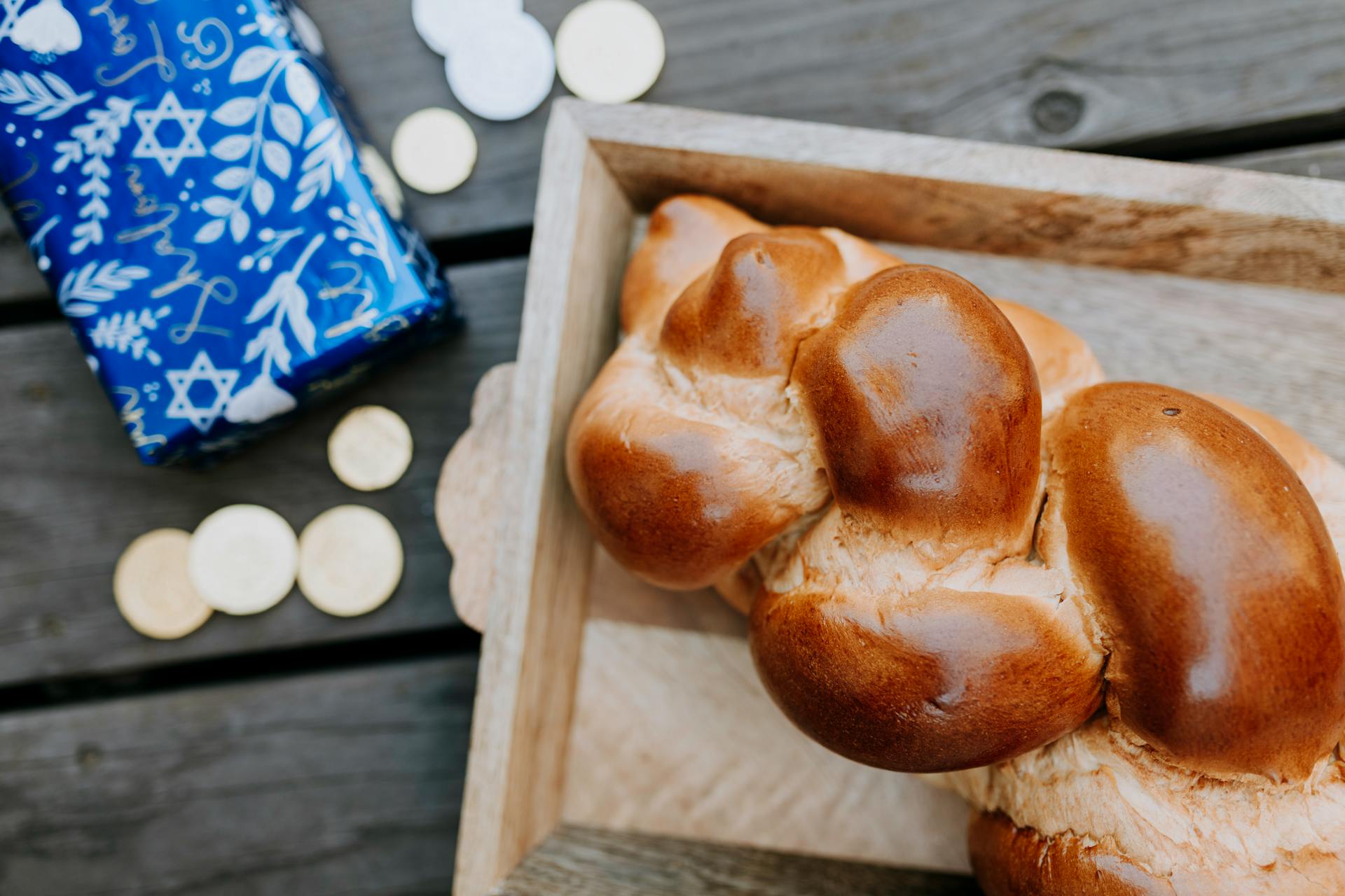 A close-up of challah bread beside Hanukkah gelt and decorative gift wrap on a wooden surface.