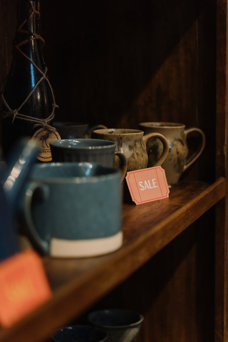 A Sale Sign Beside Cups On A Wooden Shelf