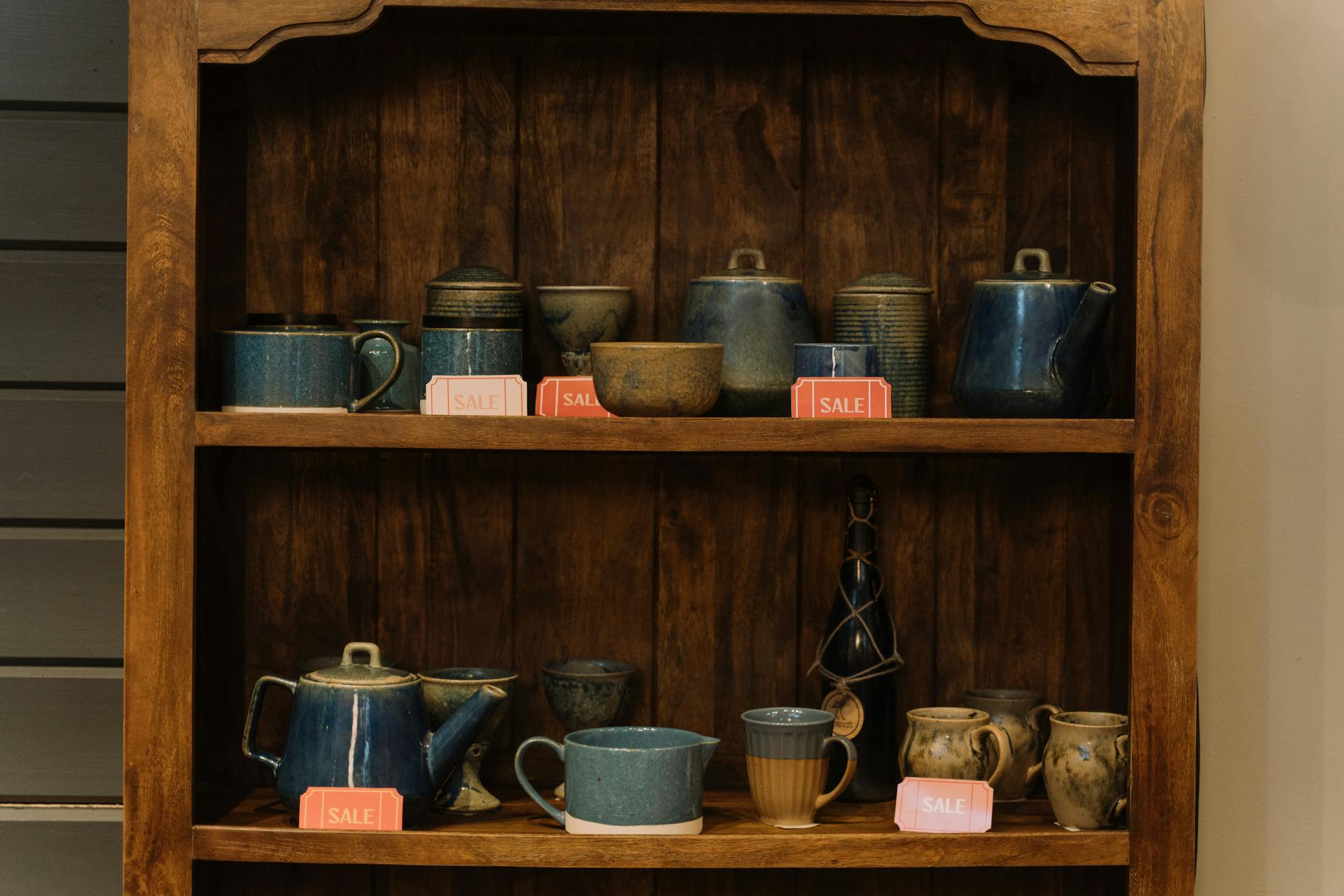 Artisan pottery cups and teapots on a wooden shelf, highlighted with sale tags.