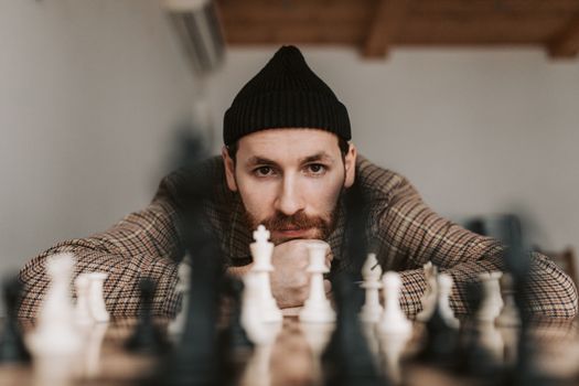 Focused man in beanie strategizing chess game indoors. Thoughtful and intense mood.