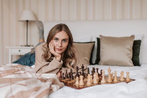 A Woman in Brown Sweater Lying on Bed Beside the Chessboard