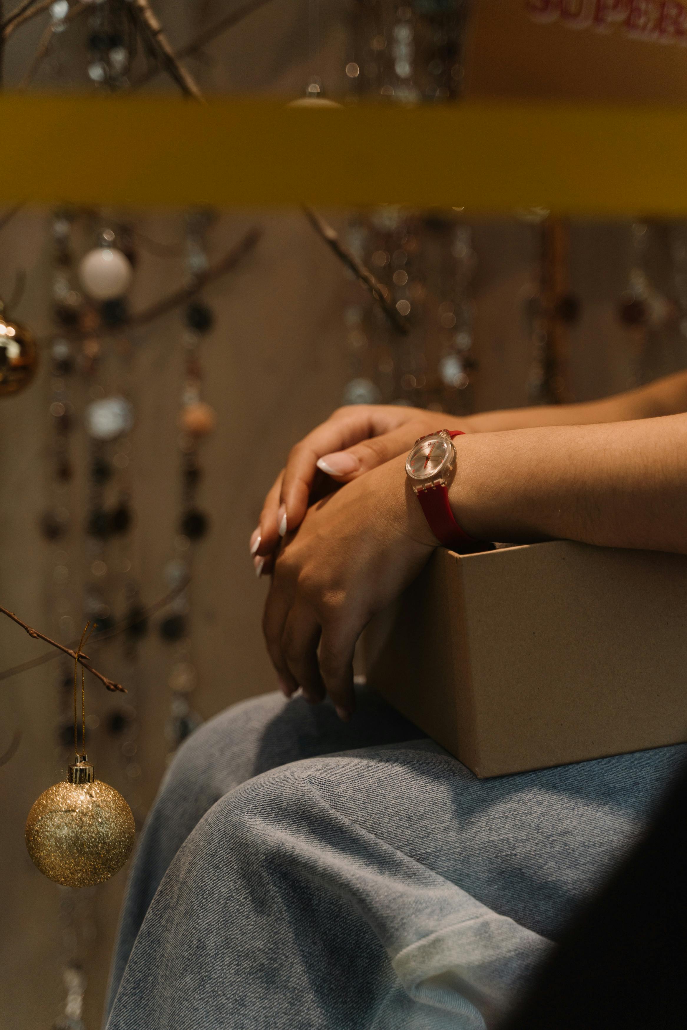 A person holding a gift box surrounded by festive Christmas baubles and decorations indoors.