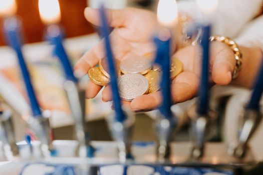 A close-up of hands holding Hanukkah gelt coins in front of a lit menorah indoors.