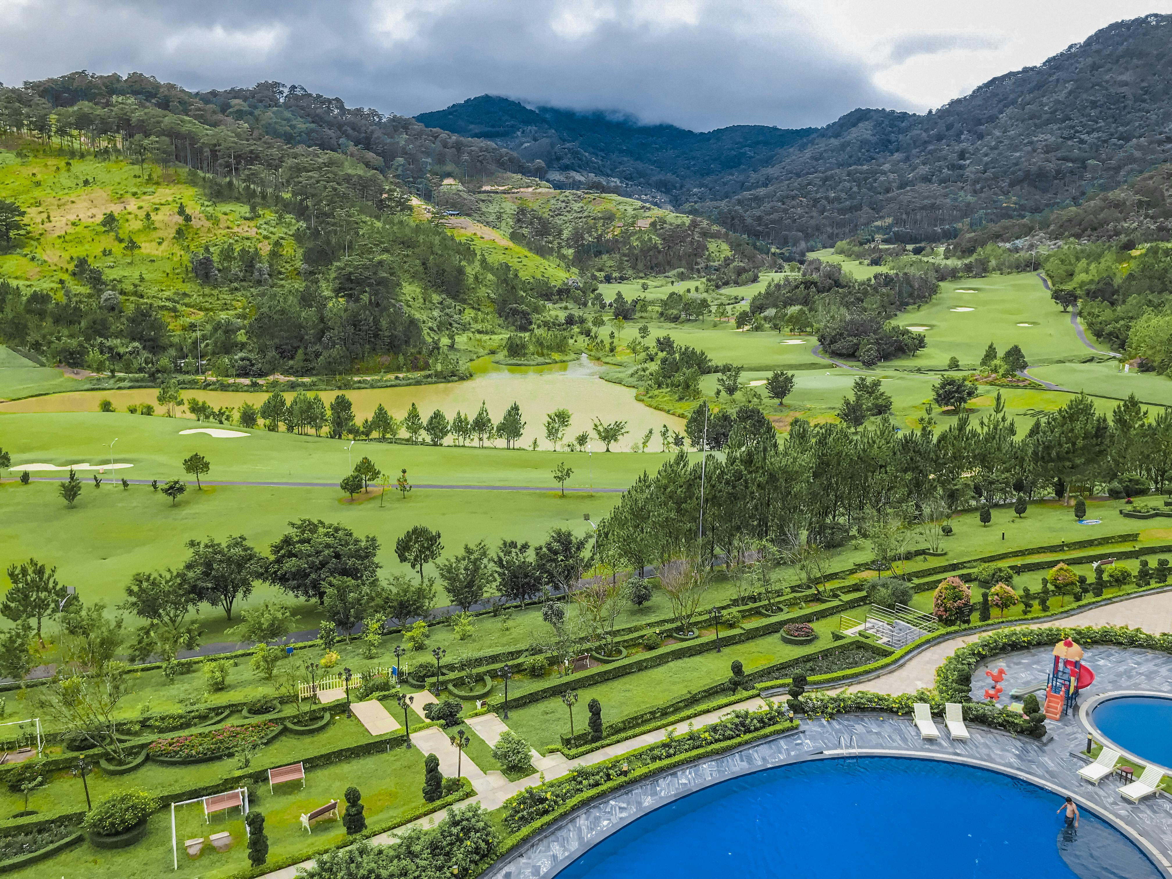 Aerial view of a lush valley with hills, trees, and a resort swimming pool under a cloudy sky.