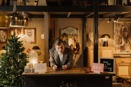 Free Man in gray suit at a shop counter with Christmas decor and sale signs, creating a festive shopping atmosphere. Stock Photo