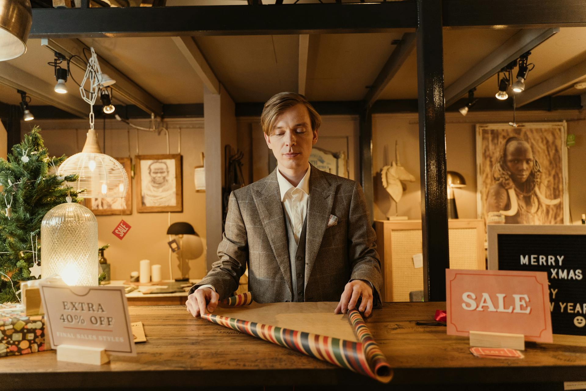 Man wrapping gifts at a retail store decorated for Christmas. Offers and sale signs visible.