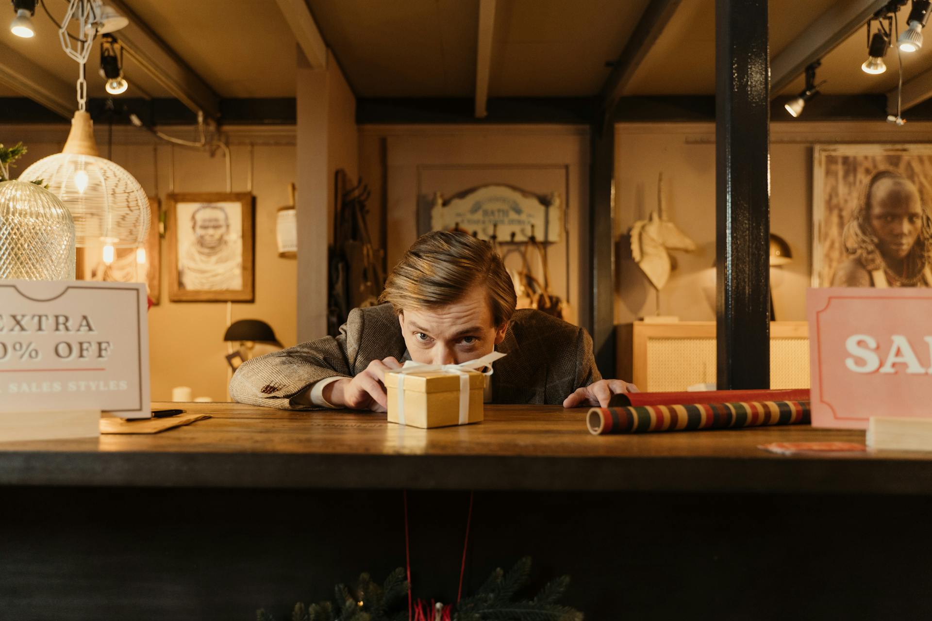 Man hiding behind gift box in a festive store setting with sale signs.