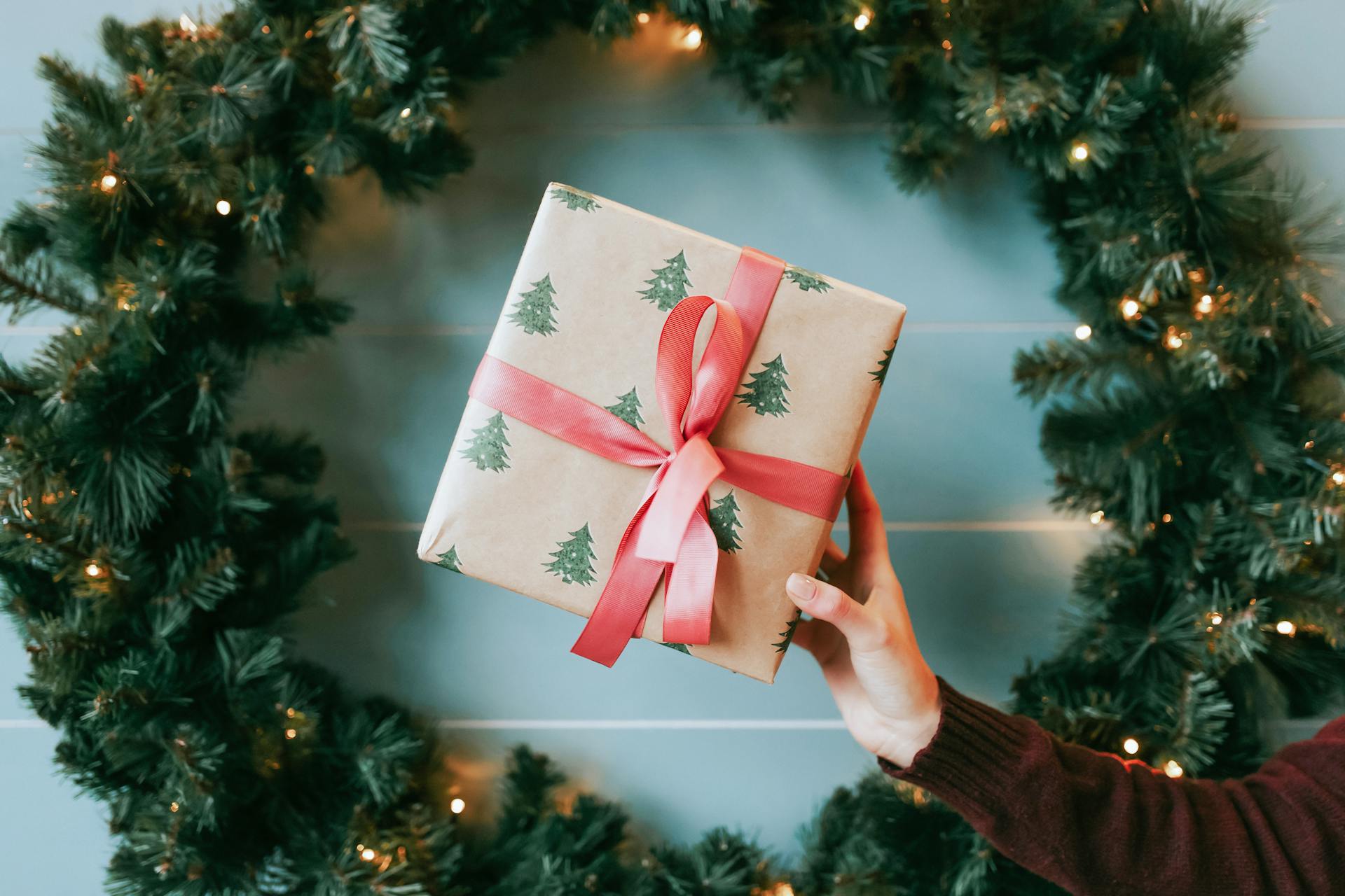 Hand holding a festive Christmas gift wrapped with tree-printed paper and red bow in front of a wreath.