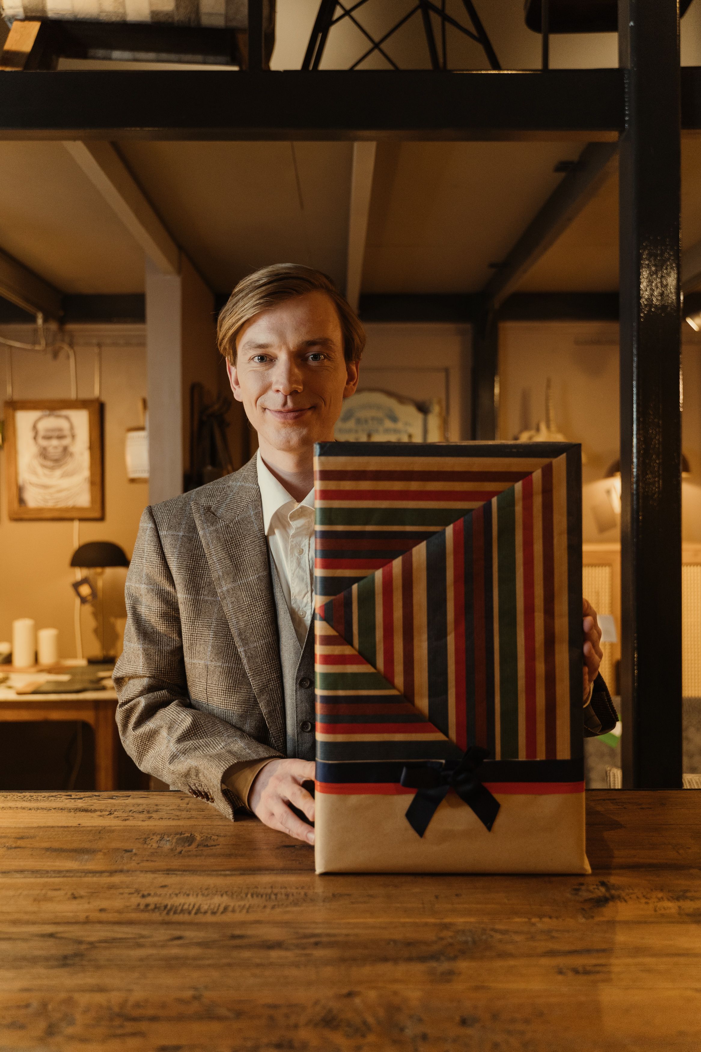 A smiling man in a suit holding a vertically striped gift box in a cozy shop setting.