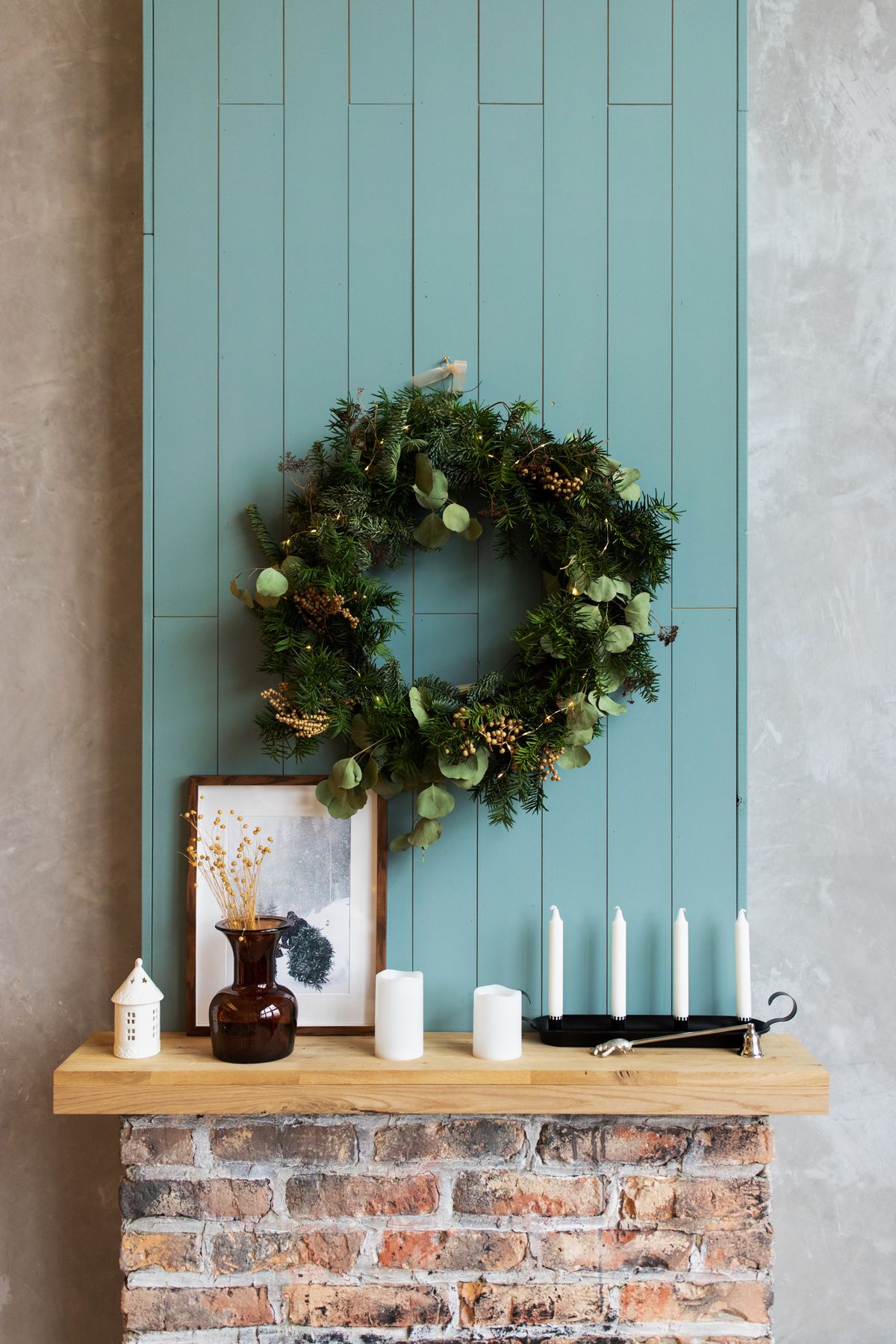 Green Christmas wreath hung on a paneled wall above a wooden mantel beam with small white candles and a dark vase