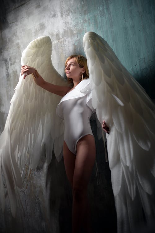 Feminine young lade wearing white bodysuit and big angle wings while standing against gray wall in studio and looking away dreamily