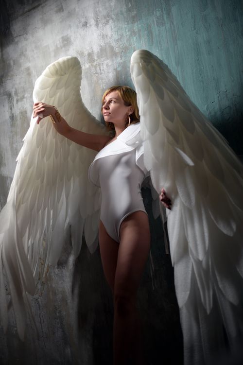 Free Feminine young lade wearing white bodysuit and big angle wings while standing against gray wall in studio and looking away dreamily Stock Photo