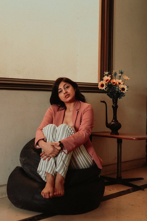 Free Full body of young pensive ethnic lady in trendy outfit sitting on bean bag chair near table with bouquet of flowers in vase and looking at camera in light apartment Stock Photo