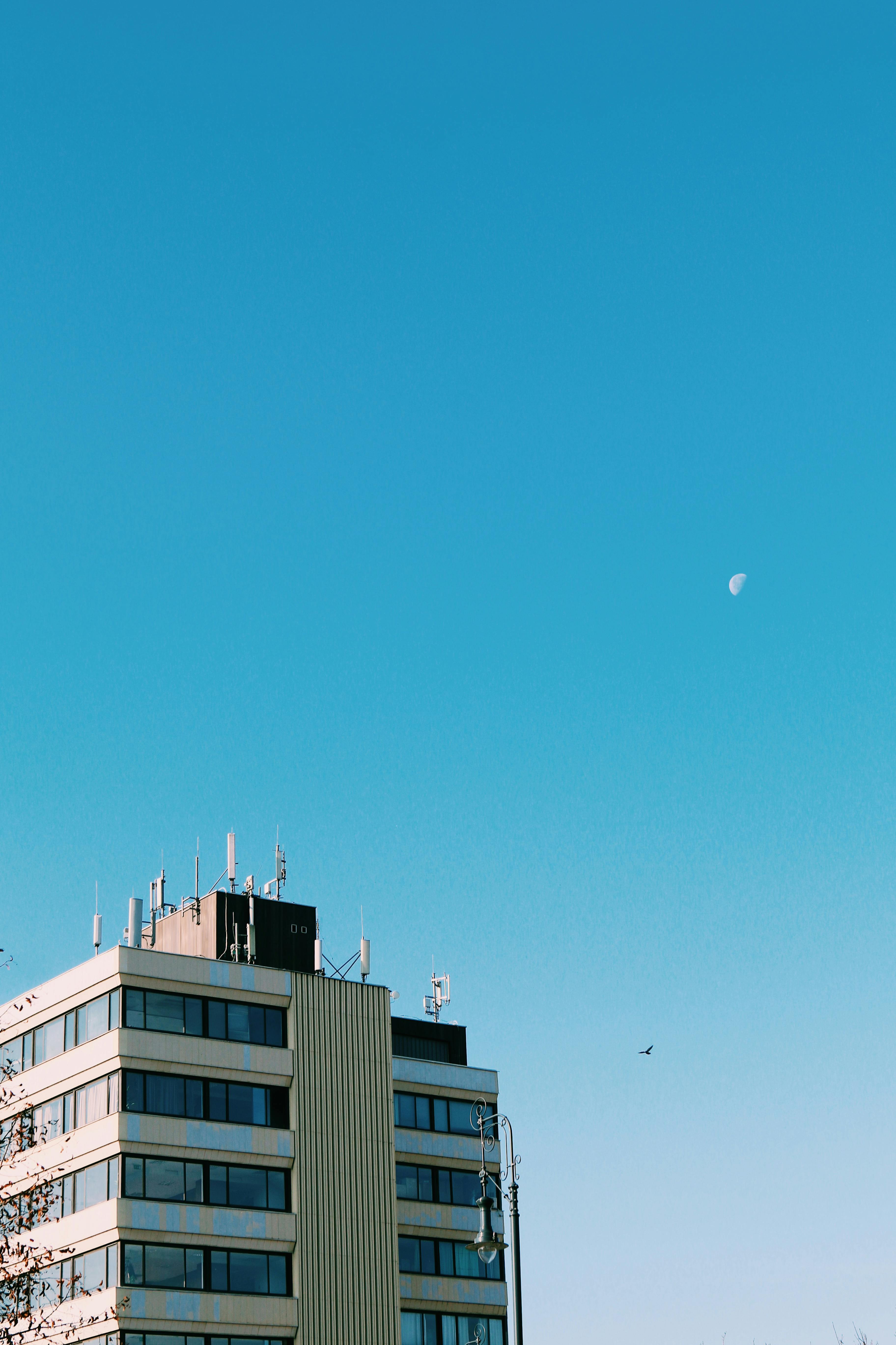 A tall modern building in Budapest with a clear blue sky and the moon visible, captured in a vertical shot.