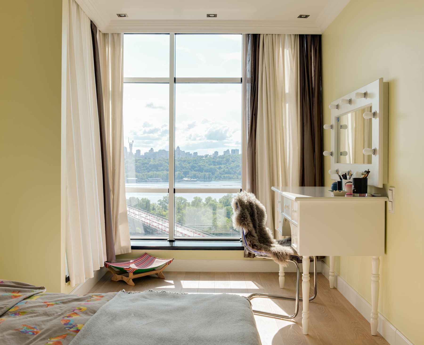 Modern bedroom with warm yellow walls, a pale upholstered bed, white bedding, dressing table and broad city window