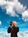 Low angle of young blond lady in trendy black outfit standing under blue cloudy sky with vintage photo camera in hands in daylight