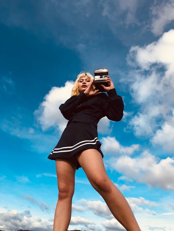 Low angle of cool Asian lady in black outfit standing with instant photo camera in hands and looking at camera under blue cloudy sky in daylight